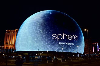 Photograph of Sphere, a large spherical entertainment venue illuminated at night with blue lighting and text reading "sphere now open." The structure is surrounded by city buildings and streetlights, highlighting its prominence and recent opening.