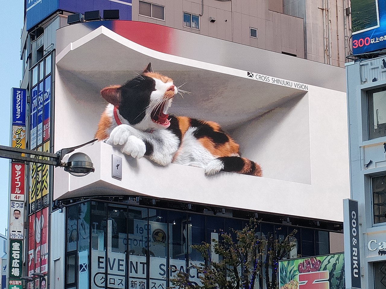 A large outdoor billboard featuring a realistic 3D-style depiction of a yawning calico cat lying in a recessed shelf. The billboard uses shading and perspective to create depth, making the cat appear to extend out from the building facade, attracting attention in a busy urban area.