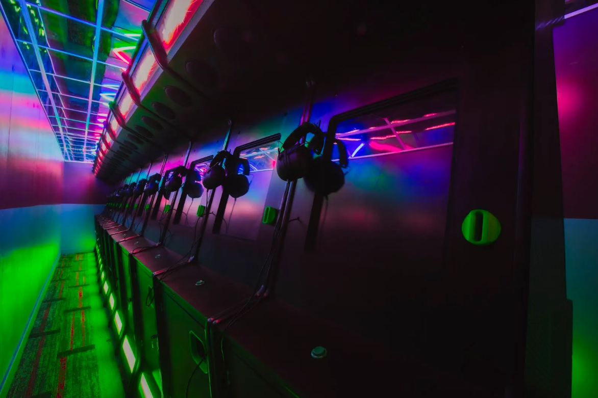 Photograph of a row of black gaming or VR stations with headsets hanging on each unit, illuminated by vibrant neon lights in green, purple, and blue. The setup suggests a futuristic gaming or virtual reality arcade environment with reflective surfaces enhancing the colorful lighting effects.