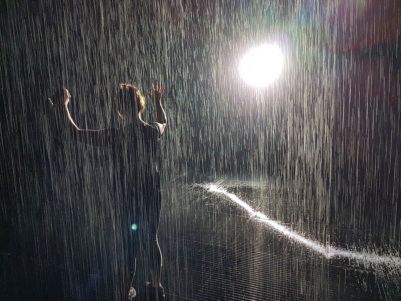 Photograph of a person standing with arms raised under heavy rainfall illuminated by a bright light source behind them. The scene highlights falling raindrops creating vertical streaks and a reflective wet surface, emphasizing motion and contrast between light and shadow.
