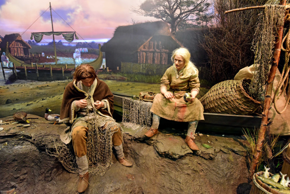 Photograph of a historical reenactment or museum display depicting two individuals dressed in medieval or Viking-era clothing engaged in fishing net repair. Scene includes rustic wooden buildings, a docked Viking-style ship with a sail, fishing nets, and baskets, set against a backdrop of a cloudy sky and barren trees.