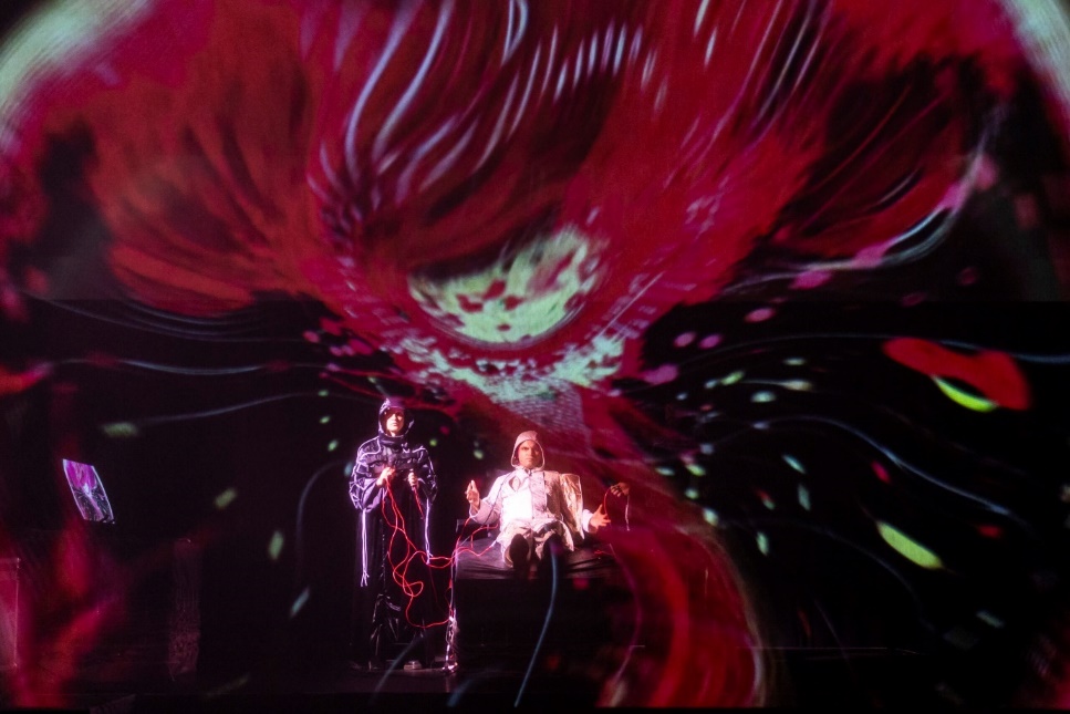 Photograph showing two individuals in a dark room with a large, vivid red and white abstract projection behind them. One person stands holding cables, while the other sits in a chair, creating a dramatic and immersive visual scene.