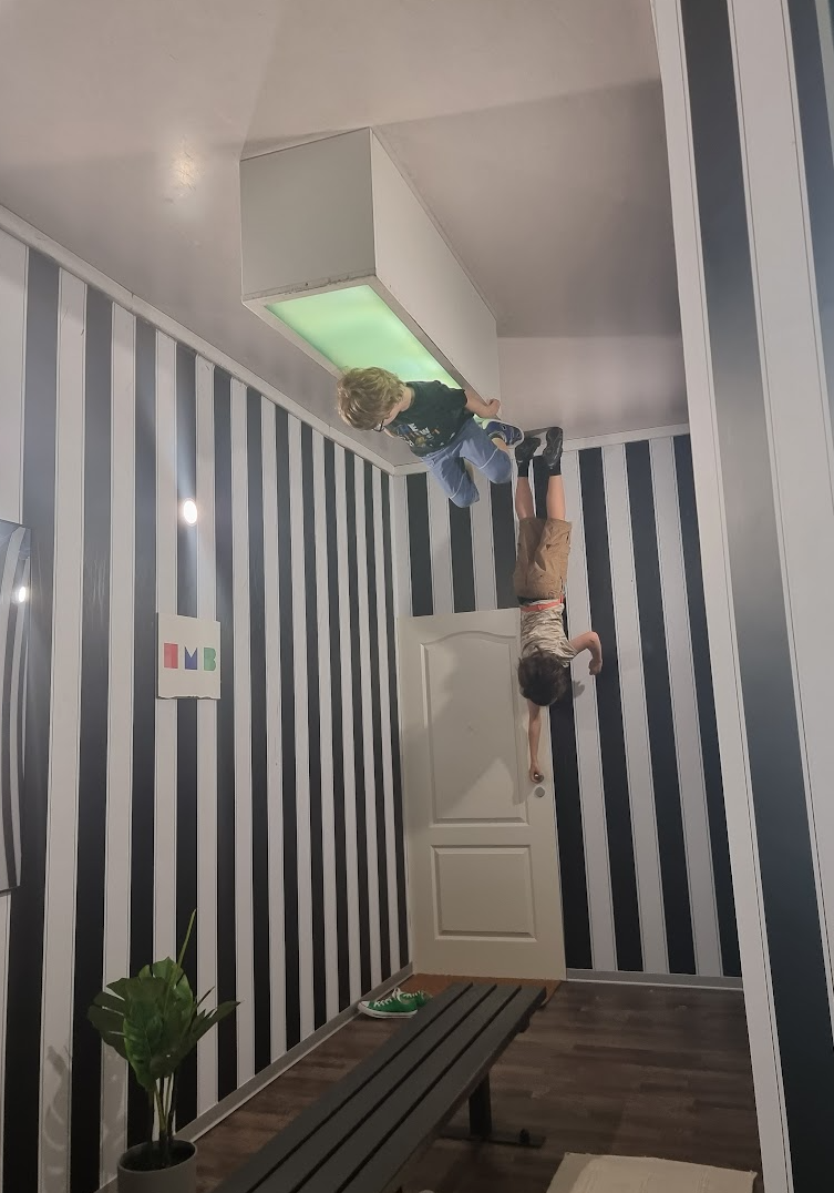 Photograph of two children appearing to hang upside down from a ceiling in a room with black and white vertical striped walls, a white door, and a bench on the floor. The room features a green-lit rectangular ceiling fixture and a small plant on the floor, creating an optical illusion of gravity reversal.