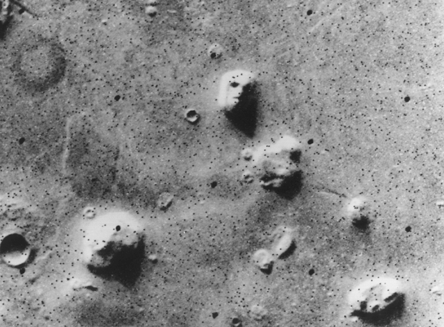 Black and white photograph showing a section of a cratered planetary surface with several raised, rounded formations resembling faces. The formations stand out due to shadows and light contrasts, highlighting unusual shapes amid a textured, pockmarked terrain.